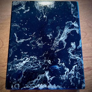 May include: A rectangular, dark blue resin box with a glossy finish. The surface features a swirling pattern of white and silver, resembling marble or a celestial nebula. The edges are a darker shade of blue.