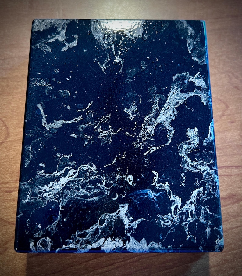 May include: A rectangular, dark blue resin box with a glossy finish. The surface features a swirling pattern of white and silver, resembling marble or a celestial nebula. The edges are a darker shade of blue.