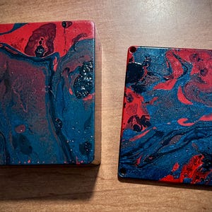 May include: Two rectangular objects with a vibrant abstract design. The design features swirling patterns of red, blue, and black. The objects appear to be made of a solid material, possibly wood or resin, and have a glossy finish. The objects are placed on a wooden surface.