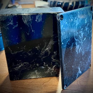 May include: A black box with a marbled white pattern, with a detached lid. The box is square and has a glossy finish. The lid is also square and has four small holes in the corners.