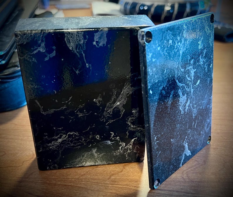 May include: A black box with a marbled white pattern, with a detached lid. The box is square and has a glossy finish. The lid is also square and has four small holes in the corners.