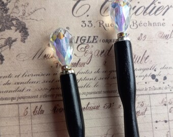 May include: Two black hair sticks with iridescent crystal-like toppers. The toppers are teardrop-shaped and catch the light. The sticks have a simple, elegant design, perfect for adding a touch of sparkle to any hairstyle. The background is a vintage paper with faded text.