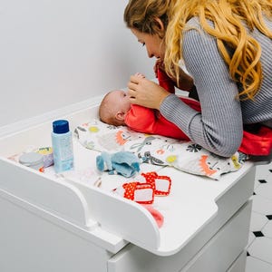 May include: A white changing table with a baby lying on a patterned changing pad. The baby is wearing a red outfit. Various baby care items are on the table, including a blue bottle and small accessories.