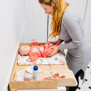 May include: A light wood baby changing table with a white cabinet base. A baby in a pink outfit lies on a patterned changing pad. Various baby care products are on the table, including diapers and lotion.