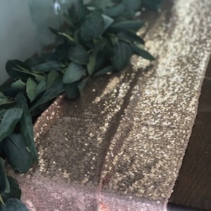 May include: A rose gold sequin table runner with a garland of green eucalyptus leaves on top.
