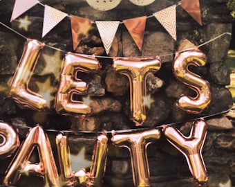 May include: A stone wall decorated with a banner of pink and brown triangles, with the words "Let's Pawty" spelled out in rose gold foil balloons. The balloons are strung together with white string.