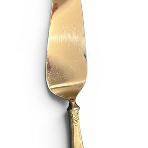 May include: A gold-colored cake server with a wide, triangular blade and a decorative handle. The handle features an ornate floral design. The server is made of metal and has a slightly worn appearance.