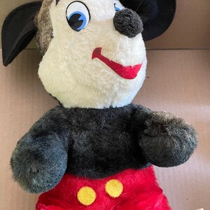 May include: A vintage plush Mickey Mouse toy with black ears, black arms and legs, red shorts with yellow buttons, and a white face with black eyes and a red mouth. The toy has a label that reads "Walt Disney Character" and "Walt Disney Productions".