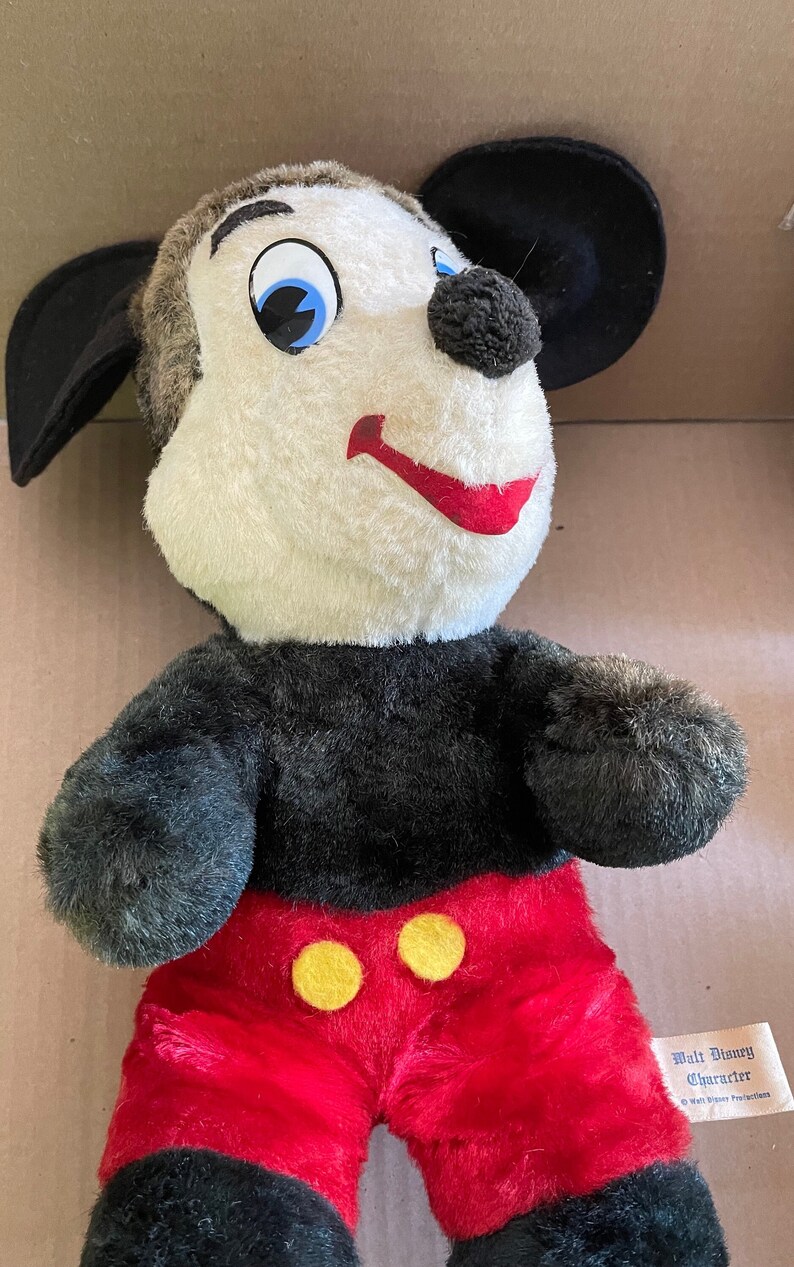 May include: A vintage plush Mickey Mouse toy with black ears, black arms and legs, red shorts with yellow buttons, and a white face with black eyes and a red mouth. The toy has a label that reads "Walt Disney Character" and "Walt Disney Productions".