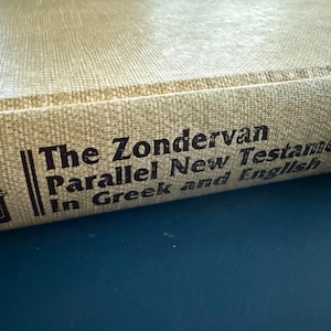 May include: A close-up of a book spine with the title "The Zondervan Parallel New Testament In Greek and English." The cover is a light tan colour with dark brown text. The book is on a dark blue surface.