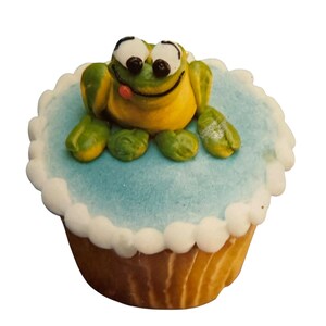 May include: A cupcake with a light blue frosting base and a white, scalloped border. A yellow and green frog figurine with a red tongue is on top of the frosting. The cupcake has a golden-brown, fluted paper cup.