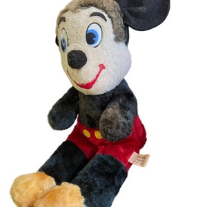 May include: Vintage plush Mickey Mouse toy with black fur, red pants, and yellow buttons. The toy has a brown head with large blue eyes and a black nose. The toy is sitting with its legs crossed and has a tag that reads "Walt Disney Character".