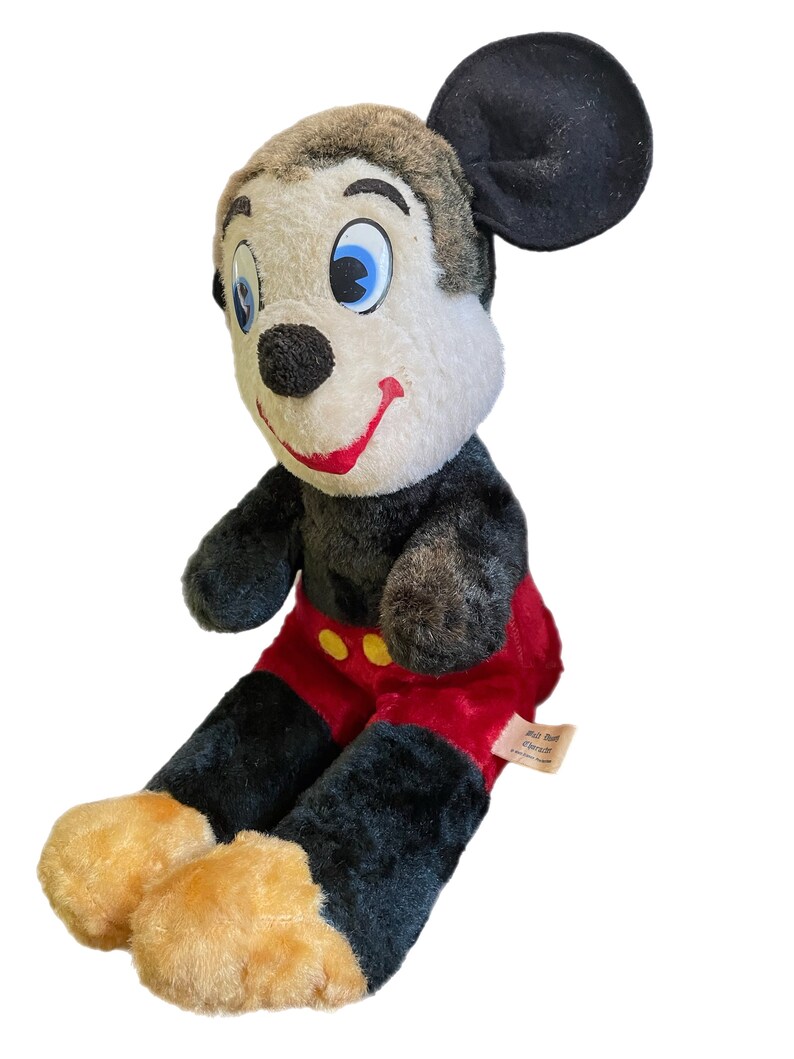 May include: Vintage plush Mickey Mouse toy with black fur, red pants, and yellow buttons. The toy has a brown head with large blue eyes and a black nose. The toy is sitting with its legs crossed and has a tag that reads "Walt Disney Character".