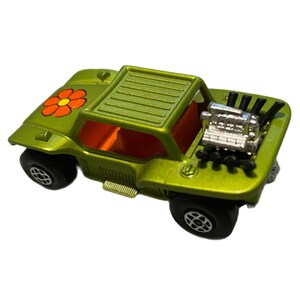 May include: A green toy dune buggy with an orange flower decal and a silver engine. The buggy has black wheels and a black roll cage.