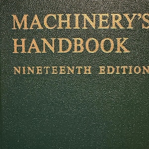 May include: Close-up of a dark green book cover with gold lettering. The title reads "MACHINERY'S HANDBOOK" above "NINETEENTH EDITION". The cover has a textured, slightly speckled appearance.