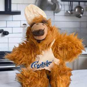 May include: A brown furry plush toy wearing a white chef's hat and a tan apron with the word "Cookin'" printed in blue.