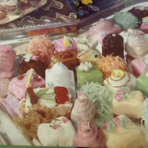 May include: A silver platter filled with an assortment of small, square-shaped petit fours. The cakes are decorated with pastel-colored icing, shredded coconut, and small sugar flowers. Some cakes feature red cherry-shaped decorations on top.