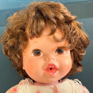 May include: A vintage baby doll with brown curly hair, brown eyes, and a pink complexion. The doll has a smiling expression with open lips and is wearing a white dress with pink accents.