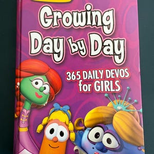 May include: A hardcover book titled "Growing Day by Day" with the VeggieTales logo. The cover features cartoon characters and the text "365 Daily Devos for Girls" on a purple background.