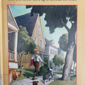 May include: A book cover illustration depicting a boy walking down a street with a red book and a dog. The book title is "The Boy on Fairfield Street: How Ted Geisel Grew Up to Become Dr. Seuss" by Kathleen Krull, with paintings by Steve Johnson and Lou Fancher.