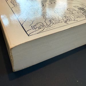 May include: A hardback book with a cream cover, displaying a black and white illustration of figures and animals. The book's pages are visible, and the cover has a glossy finish. The book is on a dark surface.