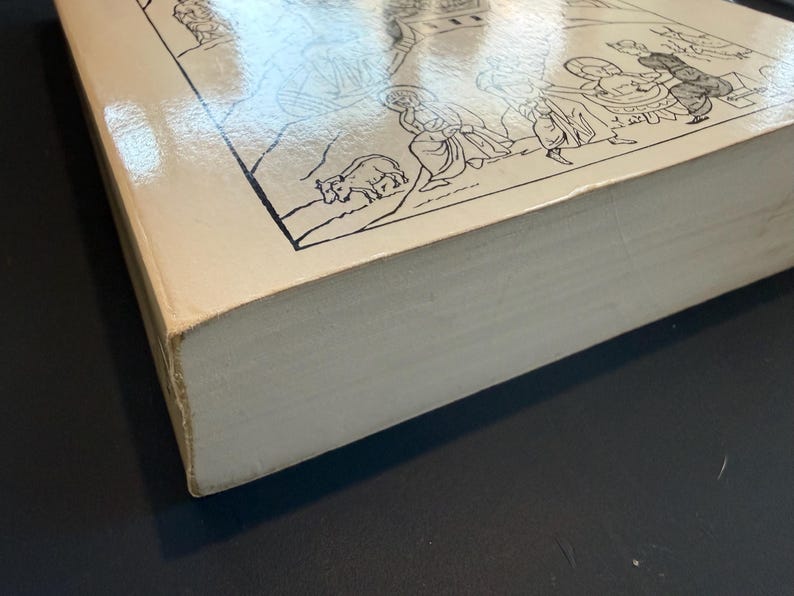 May include: A hardback book with a cream cover, displaying a black and white illustration of figures and animals. The book's pages are visible, and the cover has a glossy finish. The book is on a dark surface.