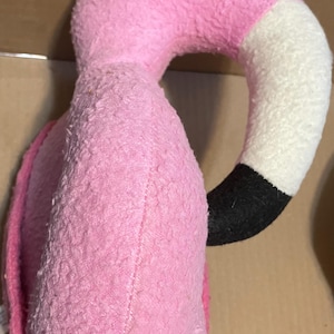 May include: Pink plush flamingo head with a white beak and black legs. The flamingo is made of a soft, fuzzy material.