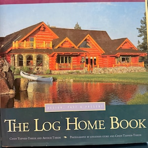 May include: A hardcover book titled The Log Home Book: Design, Past & Present by Cindy Teipner-Thiede and Arthur Thiede. The cover features a photograph of a large orange log cabin with a stone foundation, a wooden balcony, and a canoe by a pond.