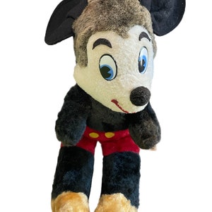 May include: A vintage plush Mickey Mouse toy with black ears, brown fur, white face, blue eyes, red shorts with yellow buttons, and tan feet.