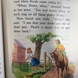 May include: A girl in a blue dress feeds a group of yellow ducklings while a girl on a brown horse rides by. The ducks are in a dirt road with a green fence and a wooden building in the background. The text in the image reads: "The ducks kept bobbing and quacking as they kept following the line of corn. They were right in the middle of the road when Sarah came riding by. "Whoa, Dusty, whoa," shouted Sarah to her pony. "You had better whoa, or we'll step on all those pretty ducks." Then Sarah saw Judy. "Hello," she said. "I see that your ducks got out of their pen. That's a smart way to get them back."