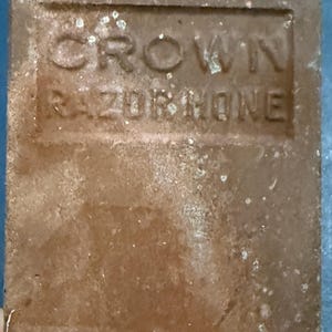 May include: A rectangular, brown razor hone with the embossed text "CROWN RAZORHONE". The surface shows signs of age and wear, with a slightly textured appearance. The hone is likely used for sharpening straight razors.
