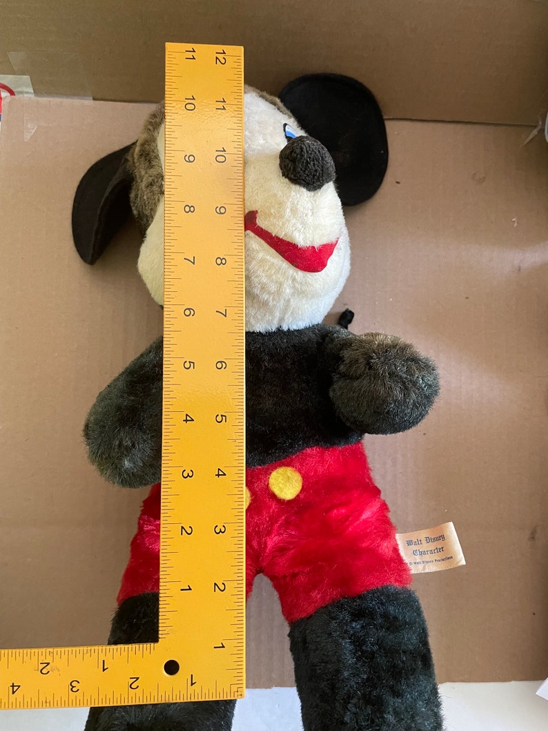 May include: A black and red plush Mickey Mouse toy with a yellow button on his shirt. The toy is 12 inches tall and has a black felt hat. The toy has a tag that reads "Walt Disney Character".
