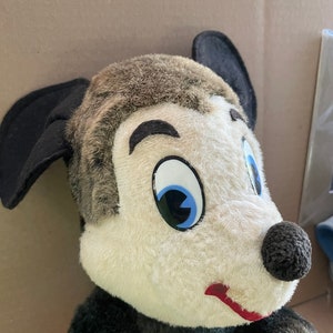 May include: A vintage plush toy of a cartoon character with black ears, a black nose, and a white face with a blue eye. The toy is wearing a black and red outfit.