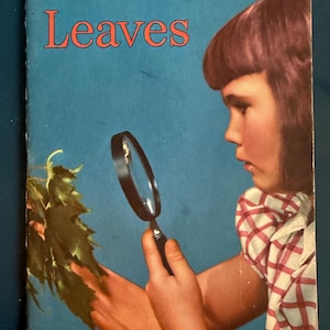 May include: A vintage children's book cover with the title "Leaves" in red letters. A young girl with pink hair is looking through a magnifying glass at a green leaf.
