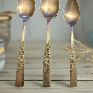 May include: Three vintage spoons with oval bowls and ornate floral designs on the handles. The spoons have a tarnished, antique gold finish, and are arranged on a light-coloured wooden surface.