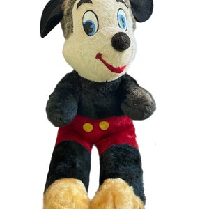 May include: A vintage plush Mickey Mouse toy with black ears, black fur, red shorts with yellow buttons, and a white face with black eyes and a red mouth.