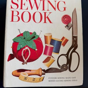 May include: A vintage book titled "Better Homes and Gardens. Sewing Book" with a red and green pincushion, spools of thread, scissors, and a thimble on the cover. The text "Custom Sewing Made Easy" and "Money-Saving Sewing Ideas" is printed at the bottom of the cover.