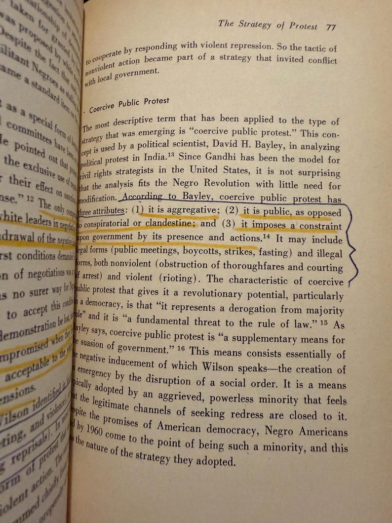 Op de afbeelding: Open boekpagina met tekst over "Coercive Public Protest" en zijn kenmerken. De tekst is in zwarte letters op verouderd, vergeeld papier. Het boek heet "The Strategy of Protest".