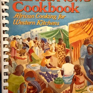 May include: The Africa News Cookbook, African Cooking for Western Kitchens, is a spiral-bound book. The cover features a vibrant illustration of an African market scene with people, food, and tents. The title is in orange and yellow.