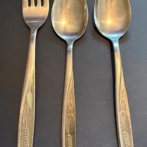 May include: A set of three silver-toned metal utensils consisting of one four-tined fork and two spoons. Each handle features an embossed wheat stalk design that extends from the base toward the neck of the utensil, with a smooth finish.