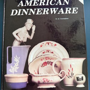 May include: A book titled "The Collector's Encyclopedia Of AMERICAN DINNERWARE" by Jo Cunningham. The cover is black with white text and features various pieces of American dinnerware, including plates, a pitcher, and a figurine.