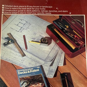 May include: A book titled Deck Plans by Ortho Books featuring construction guides, alongside a red metal toolbox, a blue architectural blueprint, a square tool, a red pencil, a yellow tape measure, and a book titled How to Design & Build Decks & Patios.
