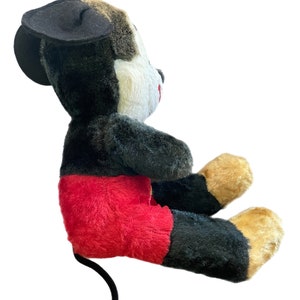 May include: A plush Mickey Mouse toy sitting with its legs crossed. The toy is black, white, and red with a black felt hat. The toy has a long black tail.