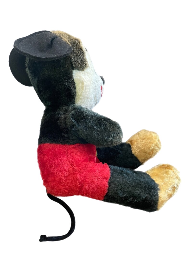 May include: A plush Mickey Mouse toy sitting with its legs crossed. The toy is black, white, and red with a black felt hat. The toy has a long black tail.