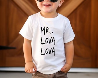 May include: A young child wearing black sunglasses, a white t-shirt with the text "MR. LOVA LOVA", brown shorts, and white sneakers. The child is smiling and standing in front of a wooden door.