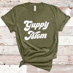 May include: Olive green t-shirt with the words "Guppy Mom" in a white, retro font. The shirt has rolled-up sleeves and a knot at the bottom. The shirt is made of a soft material.