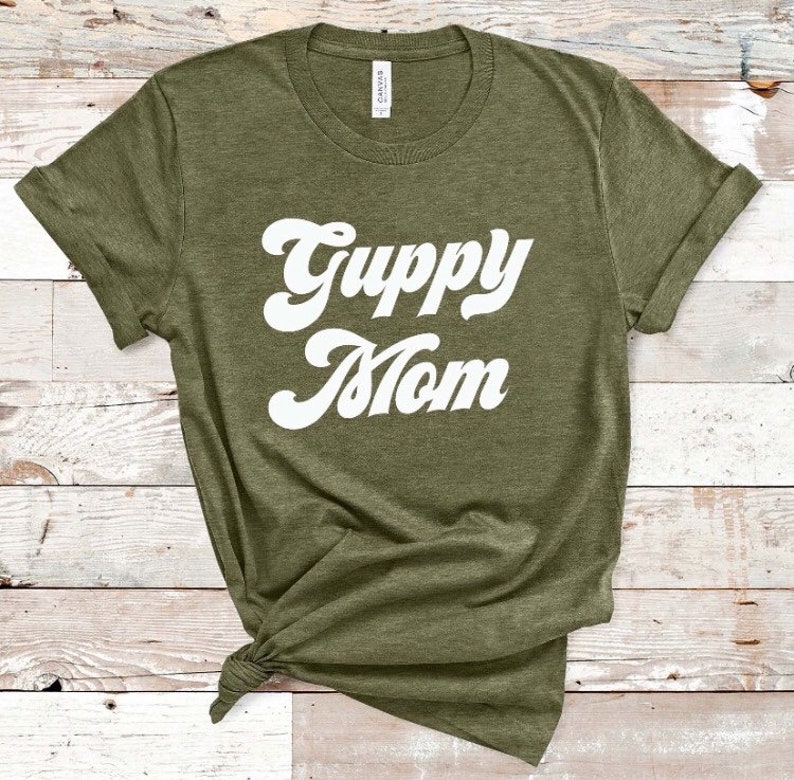 May include: Olive green t-shirt with the words "Guppy Mom" in a white, retro font. The shirt has rolled-up sleeves and a knot at the bottom. The shirt is made of a soft material.