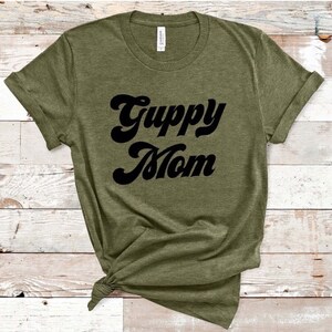 May include: Olive green t-shirt with the words "Guppy Mom" in a black, retro font. The shirt is made of a soft material and has rolled-up sleeves. The shirt is knotted at the bottom.