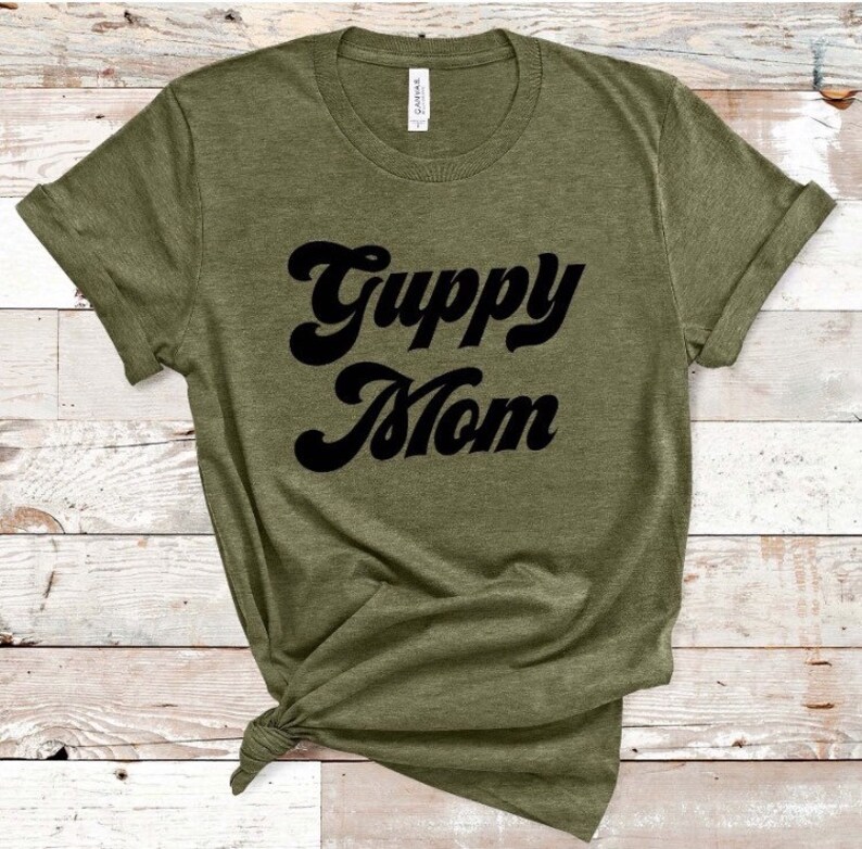 May include: Olive green t-shirt with the words "Guppy Mom" in a black, retro font. The shirt is made of a soft material and has rolled-up sleeves. The shirt is knotted at the bottom.
