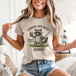 May include: Beige t-shirt with a graphic of a raccoon holding a coffee and sword, with the text "CURRENT SIDE QUEST BROUGHT TO YOU BY ADHD". The raccoon is standing on grass.